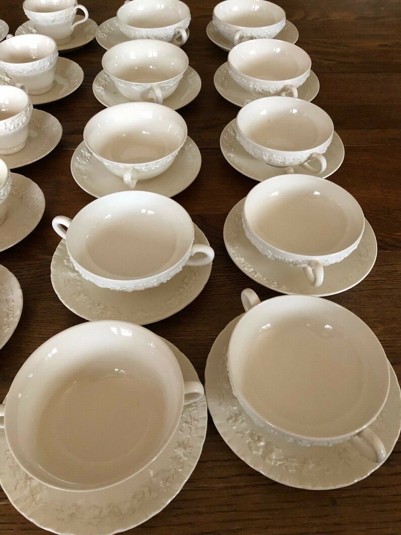 Pu&ograve; includere: Un set di 14 ciotole per zuppa in porcellana bianca con manici. Le ciotole sono disposte in file su una superficie di legno. Ogni ciotola presenta un motivo decorativo attorno al bordo.