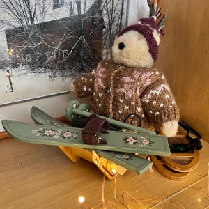 Può includere: Un orsacchiotto di peluche con un cappello di lana bordeaux e bianco con corna e un maglione abbinato. L'orso è vicino a un paio di sci di legno verdi con disegni floreali e bastoncini da sci. Una scena invernale incorniciata è sullo sfondo.