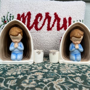 Könnte beinhalten: Zwei weiße Kerzenhalter aus Keramik mit einer Jungenfigur in jedem. Die Figuren knien mit gefalteten Händen im Gebet. Die Kerzenhalter haben ein braunes Inneres und goldene Akzente.