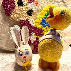Peut inclure: Deux décorations de Pâques vintage, un lapin blanc et un poussin jaune, tous deux en papier mâché et recouverts de tissu texturé. Le lapin a un visage peint avec un nez rouge et un nœud papillon jaune. Le poussin a un bec jaune et un œil noir.
