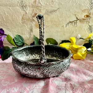 May include: A silver serving bowl with a decorative floral design and a twisted wire handle. The bowl is on a pink and white patterned surface.