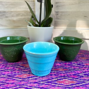 Op de afbeelding: Drie keramische bloempotten, één groen, één blauw en één wit. De groene potten hebben een decoratief ontwerp. De potten staan op een paars en roze gepatroneerd oppervlak.