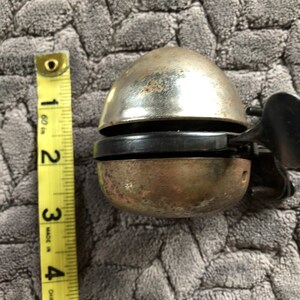 May include: A silver bicycle bell with a black rubber ring and a black metal lever. The bell is approximately 5 cm in diameter.
