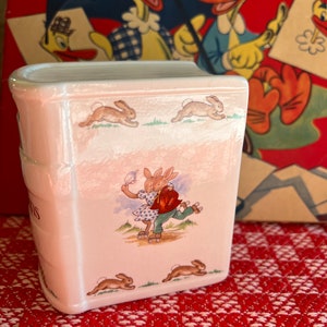 May include: A white ceramic book-shaped piggy bank with a bunny rabbit design. The bank features two bunnies roller skating and several bunnies hopping around the sides. The bank has the word "Bunnies" written on the front.