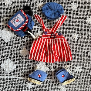 May include: A red and white striped overall outfit with a blue backpack that says "Messenger of Love" on it. The outfit also includes a blue cap and blue shoes.