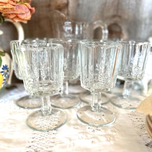 Puede incluir: Juego de seis vasos de cristal transparente con tallo y diseño texturizado. Los vasos están dispuestos sobre un mantel de encaje blanco.