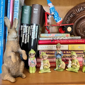 Könnte beinhalten: Eine Sammlung von Keramikfiguren von Elfen, die grüne und blaue Kleidung tragen und einen Vogel, ein Eichhörnchen und einen Welpen halten. Die Figuren sind auf einem Regal mit Büchern und einem Spielzeugclown auf einem roten Wagen angeordnet.