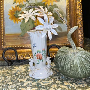 May include: A white porcelain vase with a floral design and a butterfly. The vase is decorated with a gold trim and has a pair of seahorse handles. The vase is filled with white, metallic, daisy-shaped flowers.