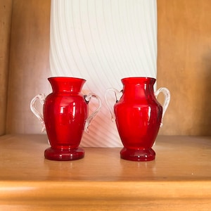 May include: Two small red glass vases with clear glass handles. The vases are sitting on a wooden shelf.