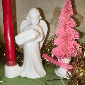 May include: A white porcelain angel figurine holding a small box, standing next to a red candle and a pink bottle brush Christmas tree. The angel is wearing a white robe and has wings.