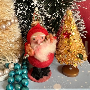 May include: A vintage Santa Claus figurine with a red felt body, white beard, and black boots. The figurine is standing on a blue and white background with a gold glitter Christmas tree.
