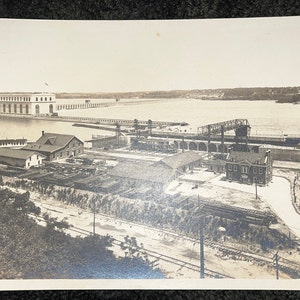 May include: A black and white vintage photograph of a large building with a long row of arches. The building is located on a body of water and there are several smaller buildings and structures in the foreground. The photo is labeled "Andrews Photo" in the bottom left corner.