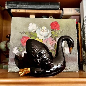 Könnte beinhalten: Eine glänzend schwarze Keramik-Schwanfigur mit goldenen Akzenten an Füßen und Schnabel. Der Schwan steht vor einem Gemälde mit Rosen in Weiß, Rosa und Rot. Die Figur steht auf einem Holzregal.