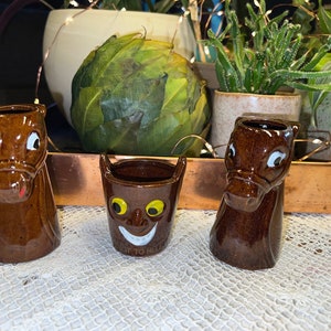 May include: Three small ceramic shot glasses with whimsical faces. The glasses are brown with white painted details. One glass is shaped like a horse's head, another is shaped like a cat's head, and the third is a round cup with a smiling face.