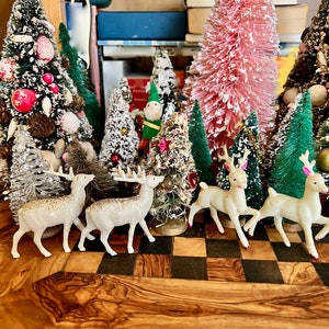 Puede incluir: Una colección de árboles de Navidad en miniatura flocados, algunos con flocado blanco, otros con flocado rosa y otros con flocado verde. También hay cuatro figuritas de ciervos blancos con narices rojas y una pequeña figurita de elfo de pie frente a los árboles. Los árboles están dispuestos sobre una superficie a cuadros.