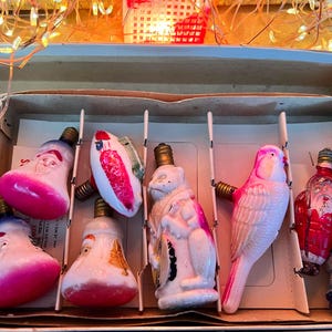May include: A collection of seven vintage figural Christmas light bulbs in a cardboard box. The set includes two Santa Claus heads with pink bases, a red and white striped ornament, a white monkey, a pink bird, a red Santa, and a blue mushroom.