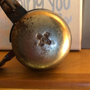 May include: A vintage silver bicycle bell with a worn finish and a cross-shaped design in the centre. The bell is attached to a black metal bracket.
