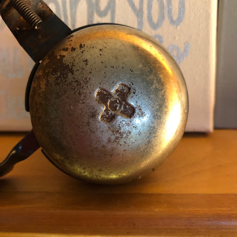 May include: A vintage silver bicycle bell with a worn finish and a cross-shaped design in the centre. The bell is attached to a black metal bracket.