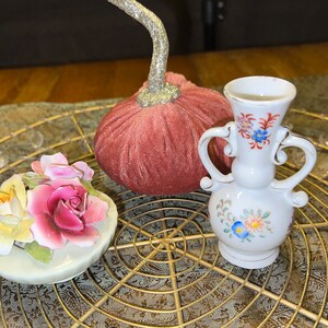 Può includere: Un set di tre oggetti decorativi: una zucca di velluto rosa, un vaso di porcellana bianca con motivi floreali e un bocciolo di rosa di porcellana bianca con una rosa gialla e foglie verdi.