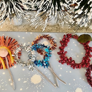 Puede incluir: Cuatro adornos navideños hechos a mano. Uno es una corona de bayas rojas, otro es una corona de oropel rojo y naranja, otro es una corona de oropel plateado y azul, y el último es una corona de oropel rojo y naranja con un centro de hilo amarillo.