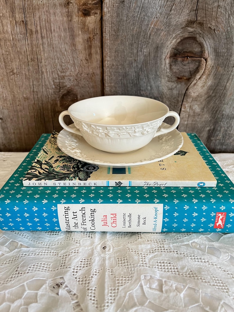 Pu&ograve; includere: Una ciotola per zuppa in ceramica bianca con un motivo floreale si trova su un piattino bianco. La ciotola e il piattino sono impilati su due libri. Il libro superiore &egrave; intitolato "La Perla" di John Steinbeck. Il libro inferiore &egrave; intitolato "Padroneggiare l'arte della cucina francese" di Julia Child, Louisette Bertholle e Simone Beck.