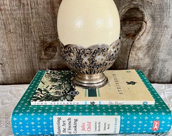 May include: A large, white ostrich egg sits in a silver egg cup on top of two vintage books. The books are titled "The Pearl" and "Mastering the Art of French Cooking" by John Steinbeck and Julia Child.