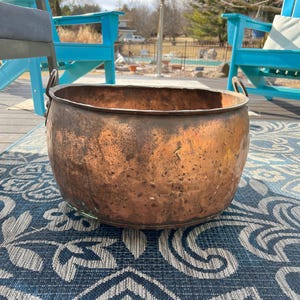 Puede incluir: Un macetero grande de cobre con dos asas se encuentra sobre una alfombra de patrón azul y gris. El macetero está ligeramente abollonado y tiene un aspecto rústico.