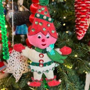 May include: A felt Christmas ornament of an elf wearing a red hat with green sequins, a white shirt with green sequins, and red mittens. The elf has a pink face with black eyes and a red mouth.