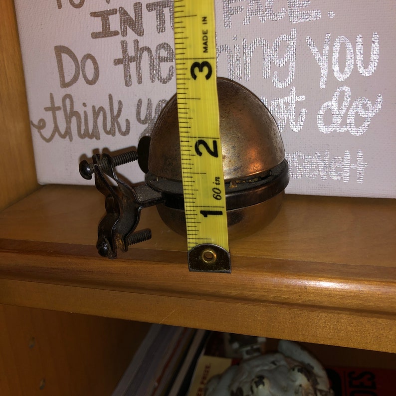 May include: A brass bicycle bell with a black metal bracket. The bell is 2 inches in diameter and is measured with a yellow tape measure.