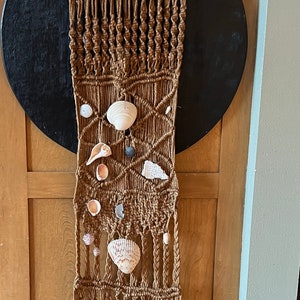 May include: A brown macrame wall hanging with a variety of seashells attached to it. The wall hanging is made of natural fibers and has a fringed bottom.