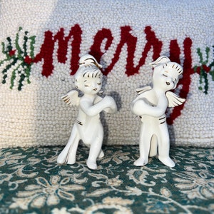 Peut inclure: Deux figurines d'anges en porcelaine blanche avec des accents dorés se tiennent devant un coussin blanc avec le mot "Merry" brodé en rouge. Le coussin est orné de branches de houx vertes de chaque côté du mot.
