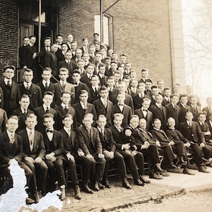 May include: A black and white photograph of a large group of men, likely students, standing in front of a brick building. The men are wearing suits and ties, and some are holding hats. The photo appears to be from the early 1900s.