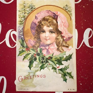 Bonita postal victoriana de saludos navideños con una chica en un gorro rosa, rizos de plátano, acebo y altos pinos nevados c.1908 NY