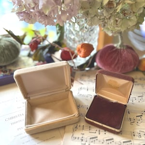 May include: Two open jewelry boxes, one beige and one burgundy, are sitting on a sheet of music. The boxes are empty and have a velvet interior.