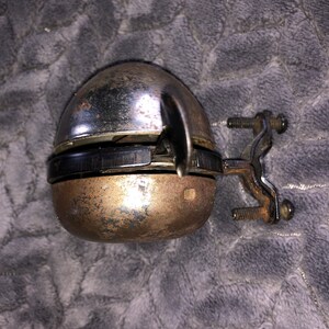 May include: A vintage bicycle bell with a chrome finish and a black rubber ring. The bell is attached to a metal bracket with two screws.