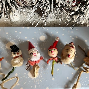 May include: Six vintage Christmas ornaments, including a Santa Claus, an elf, and four snowmen. The ornaments are made of cotton and have painted faces. They are attached to wire stems.