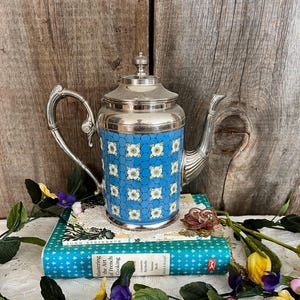Könnte beinhalten: Eine silberne Teekanne mit einem blauen und weißen Blumenmuster. Die Teekanne steht auf einem Stapel Bücher mit einem blauen Einband.