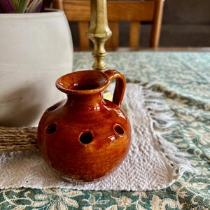 May include: A small, brown ceramic vase with a handle and six holes for flowers. The vase is sitting on a white and green patterned tablecloth.