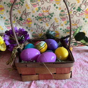 May include: A brown wicker basket filled with colorful plastic Easter eggs. The basket has a brown handle and is sitting on a pink tablecloth with a floral pattern. The eggs are purple, blue, yellow, and one has a picture of a bunny on it.