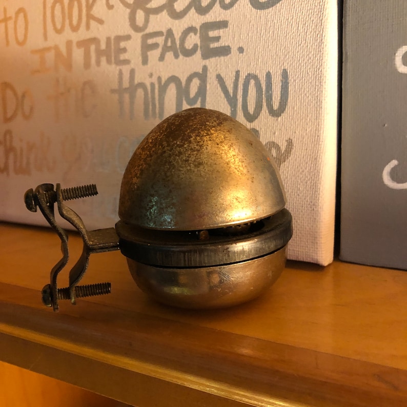 May include: A vintage, silver-toned bicycle bell with a black band around the middle. The bell is mounted on a metal bracket with two screws.