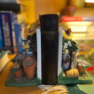 May include: A brown leather-bound book titled "Holy Bible Scofield Reference Edition" is held upright by a pair of decorative bookends. The bookends are made of ceramic and feature a garden theme with flowers, pots, and a watering can.