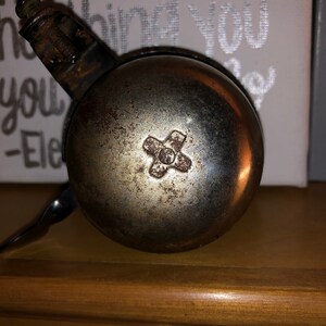 May include: A silver bicycle bell with a rusty cross-shaped design in the centre. The bell is attached to a black metal bracket.