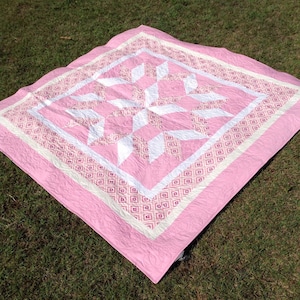 Puede incluir: Una colcha rosa y blanca con un patr&oacute;n geom&eacute;trico. La colcha est&aacute; hecha de tela de algod&oacute;n y tiene un dise&ntilde;o acolchado.