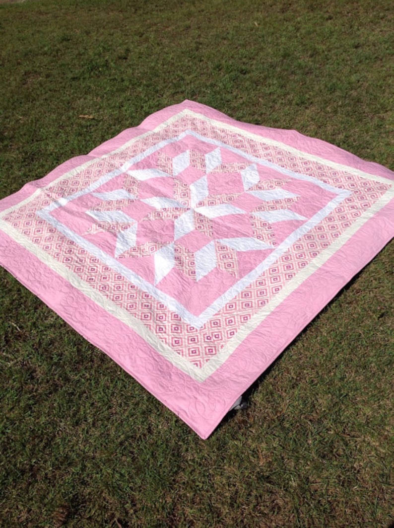 Puede incluir: Una colcha rosa y blanca con un patr&oacute;n geom&eacute;trico. La colcha est&aacute; hecha de tela de algod&oacute;n y tiene un dise&ntilde;o acolchado.