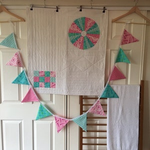 May include: A white quilt with a pink and green circular design in the centre and a patchwork square design in the bottom left corner. A white towel with a pink and green trim is hanging on a wooden rack. A string of pink, green and white triangular flags is hanging above the quilt and towel.