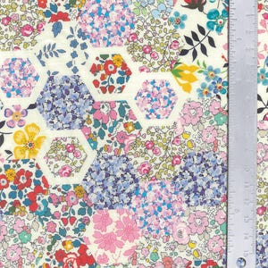 May include: A patterned fabric with a floral design. The fabric displays a variety of colorful flowers in shades of pink, blue, red, and yellow, arranged in hexagonal shapes. A metal ruler is placed on the right side of the fabric, showing centimetre measurements.