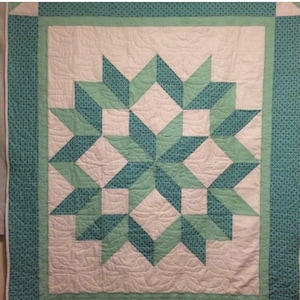 May include: A handmade quilt with a central starburst design in teal and light green. The star pattern is set against a cream background, with a teal patterned border. The quilt has a quilted texture, with a decorative stitch pattern.