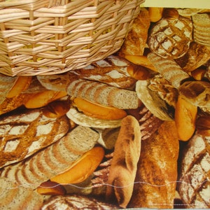 May include: A woven wicker basket rests on a placemat featuring a bread-themed pattern. The placemat showcases various types of bread, including baguettes and loaves, in shades of brown and tan. The basket is made of light brown woven wicker.