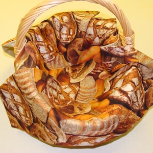 May include: A woven wicker basket with a curved handle. The interior is lined with fabric featuring a print of various types of bread in shades of brown and tan. The basket is empty.