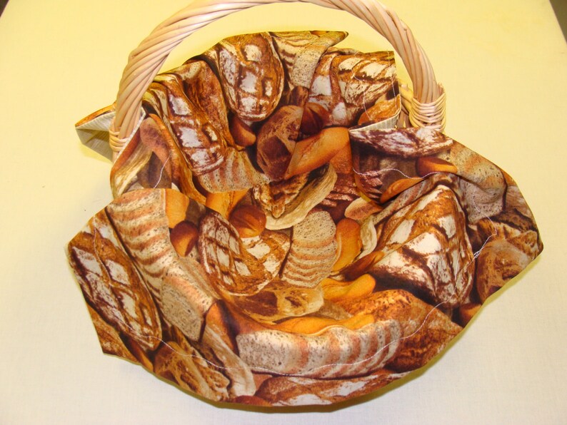 May include: A woven wicker basket with a curved handle. The interior is lined with fabric featuring a print of various types of bread in shades of brown and tan. The basket is empty.